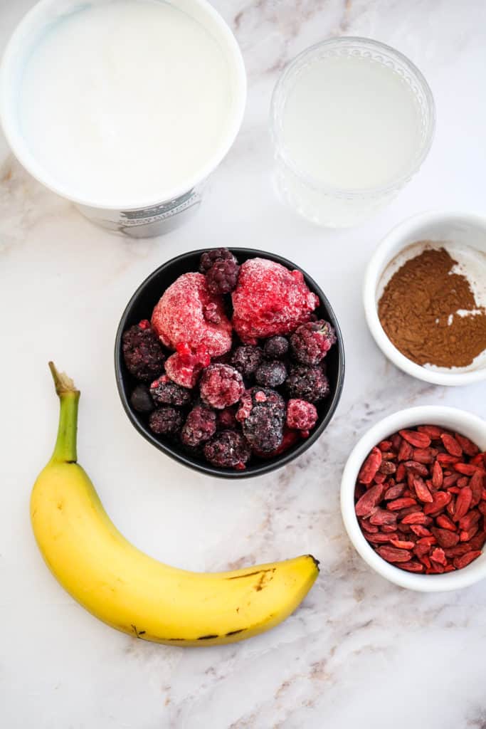 Frozen mixed berries, banana, goji berries, cinnamon, coconut water, and So Delicious Coconutmilk Yogurt Alternative on a white countertop.