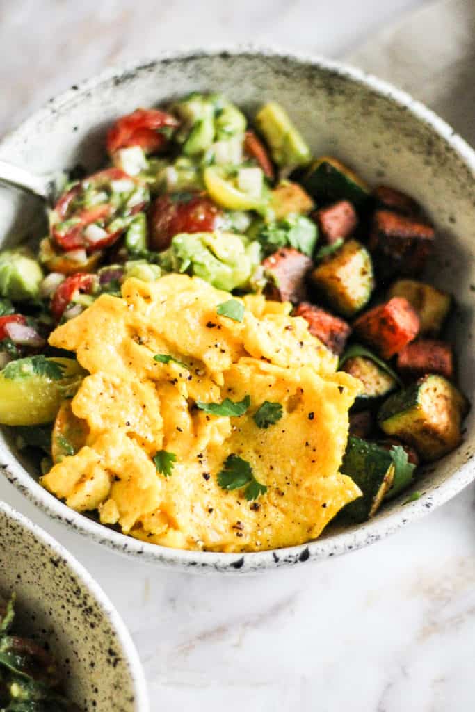 Scrambled egg in a white speckled ceramic bowl with sautéed vegetables and avocado salad.