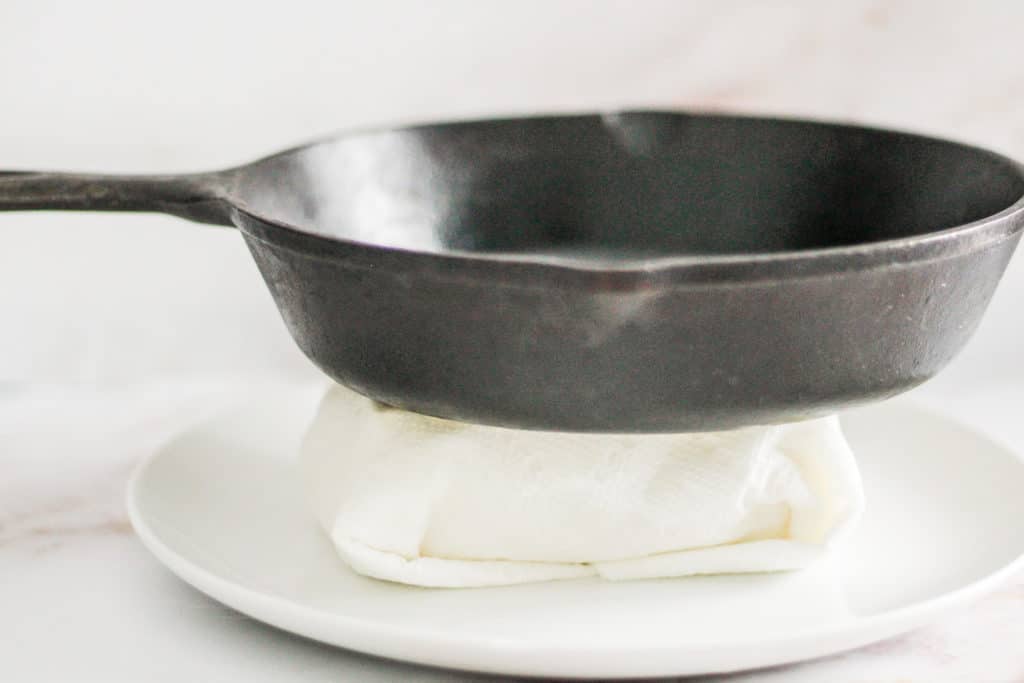 Tofu wrapped in paper towel on a white plate with a cast-iron skillet on top of it.
