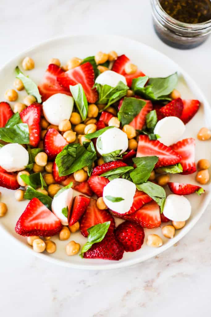 Sliced strawberries, mozzarella, chickpeas, and basil on a white plate.