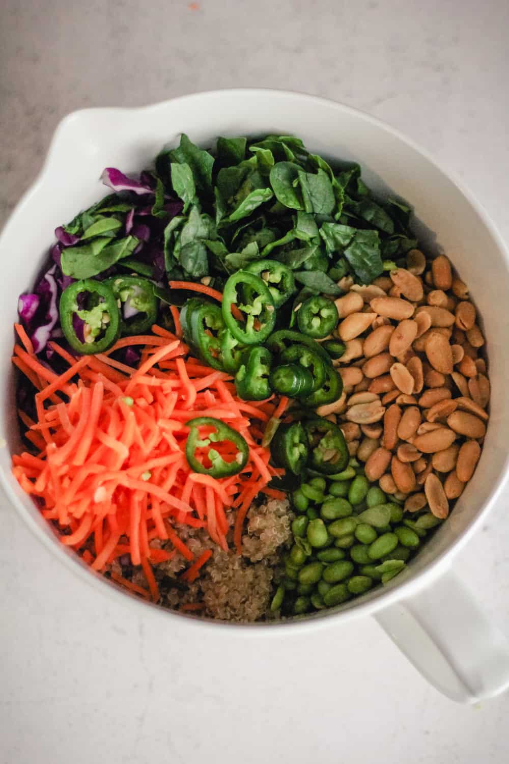 Salad ingredients in a large mixing bowl, including spinach, cabbage, carrots, peanuts, quinoa, edamame, and jalapeño. 