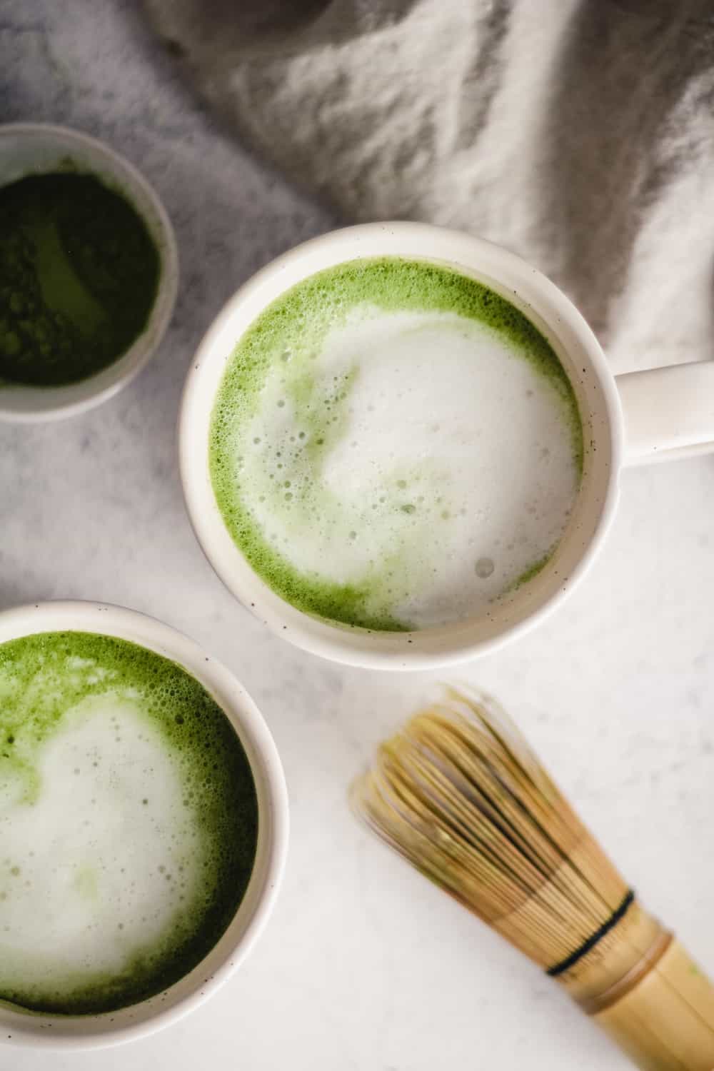 Overhead image of vanilla matcha lattes in white mugs with a bamboo matcha whisk.