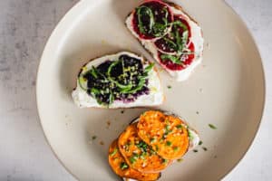 Horizontal image of yogurt toast on a beige plate. Toast is topped with sweet potato, wild blueberries, and blood orange.
