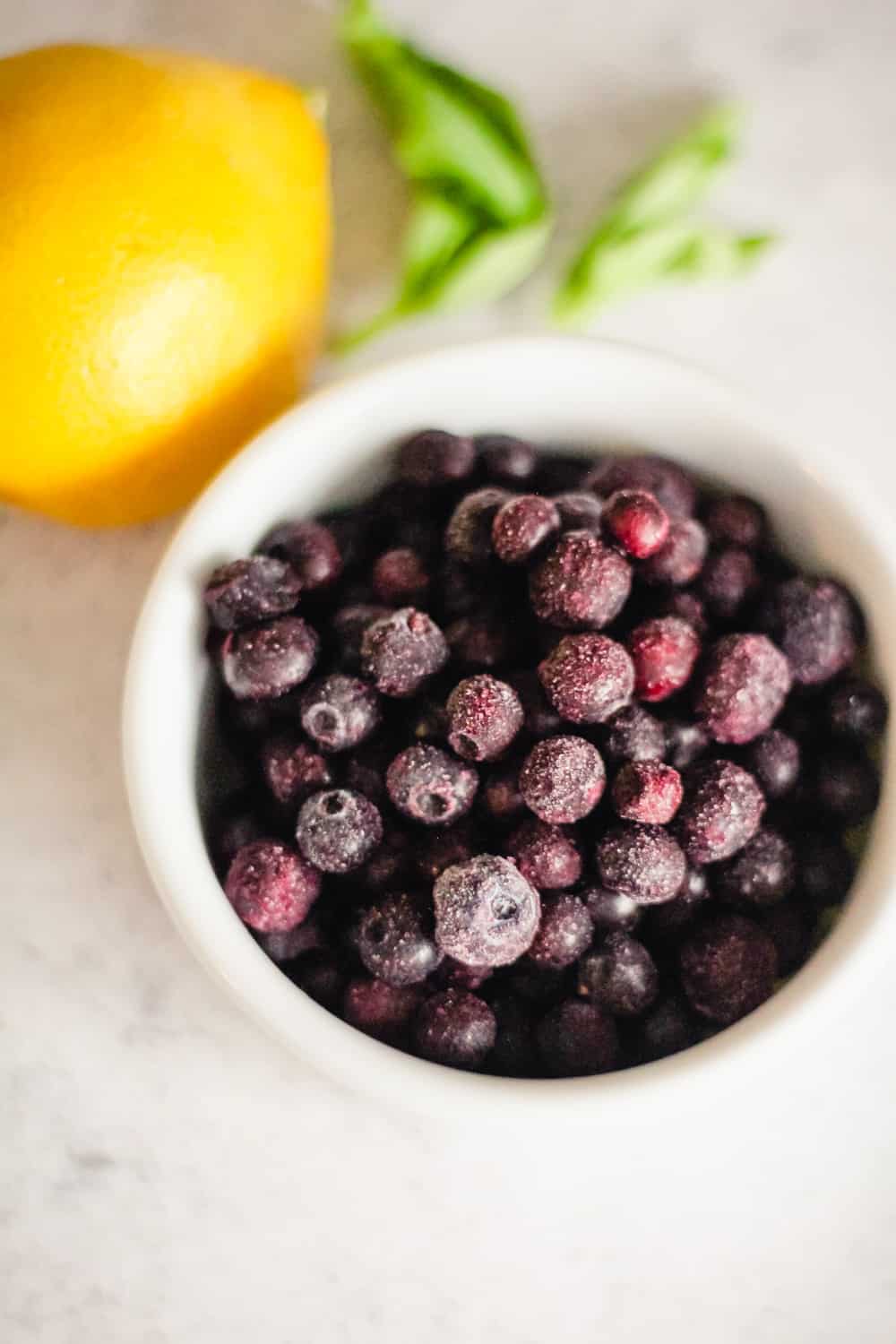 Frozen wild blueberries in a bowl with lemon and basil behind it. 