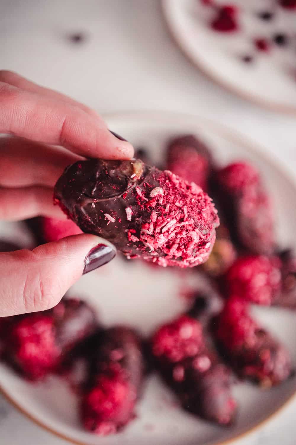 Hand holding a chocolate date with coconut and raspberry topping.