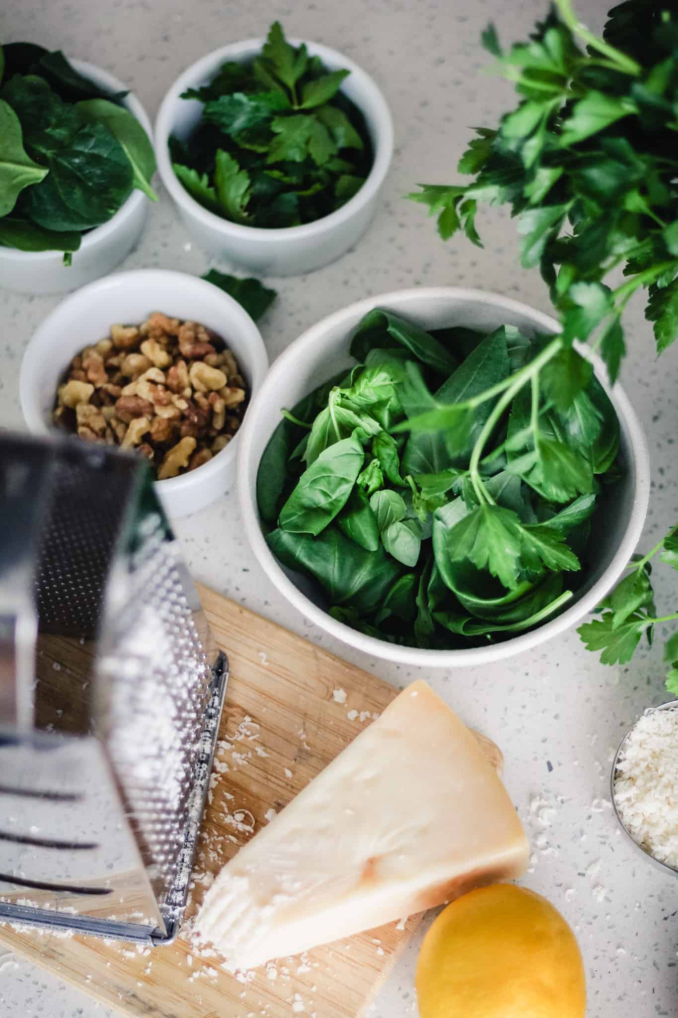 A wedge of Parmesan cheese on a cutting board with a grater next to lemon and bowls of basil, walnuts, spinach and parsley.