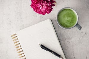 Horizontal flat-lay of a notebook with a cup of matcha and a pink flower.