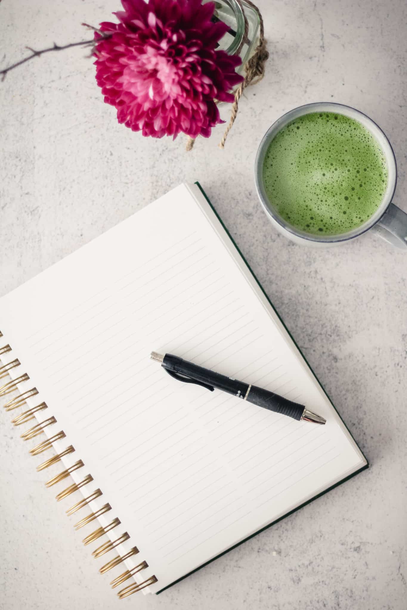 Flat-lay of a notebook with a cup of matcha and a pink flower.