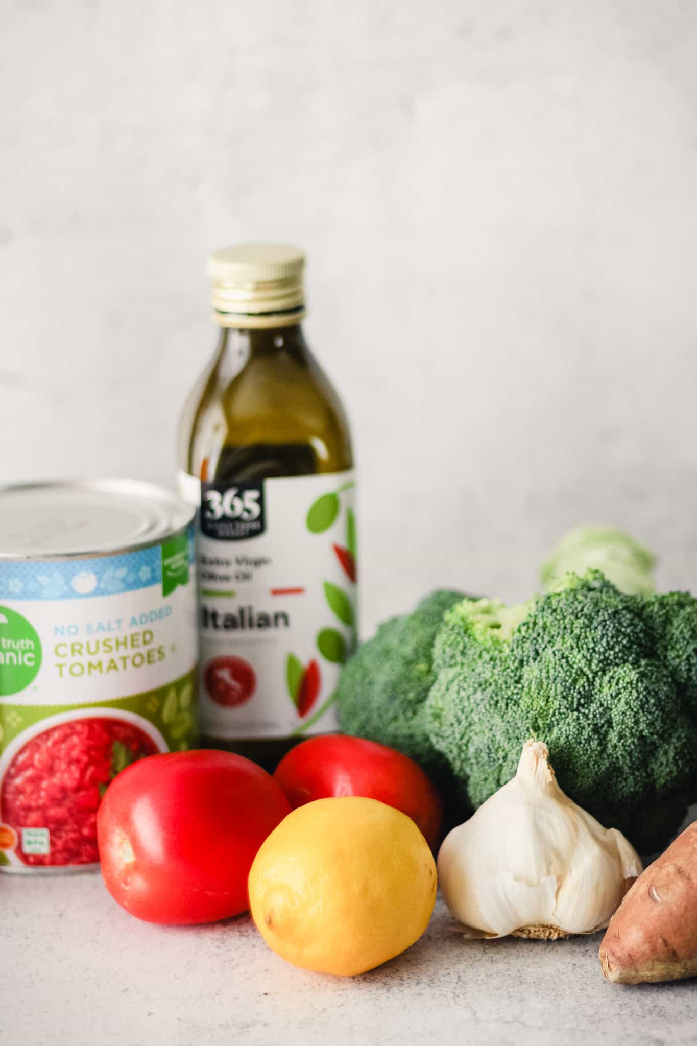 Vegretables, canned tomatoes, and a bottle of olive oil against a light background.