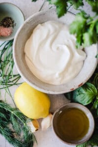 Flat-lay image of yogurt alternative, fresh herbs, olive oil, lemon, garlic, salt and pepper.