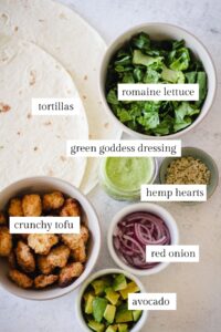 Flat-lay omage with labeled ingredients: tortillas, romaine lettuce, green goddess dressing, hemp hearts, crunchy tofu, red onion, and avocado.