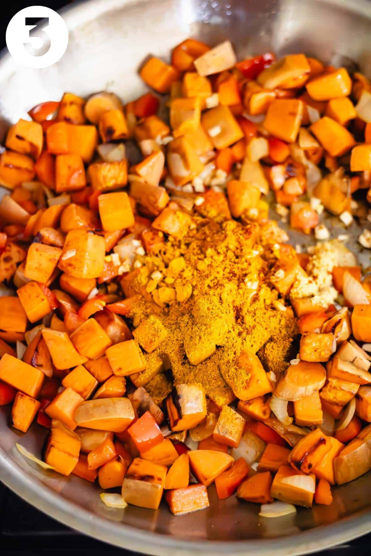 Ground spices, garlic, and ginger are sitting on top of a skillet with cubed sweet potato and diced onion and red bell pepper. The photo is labeled with a number "3."