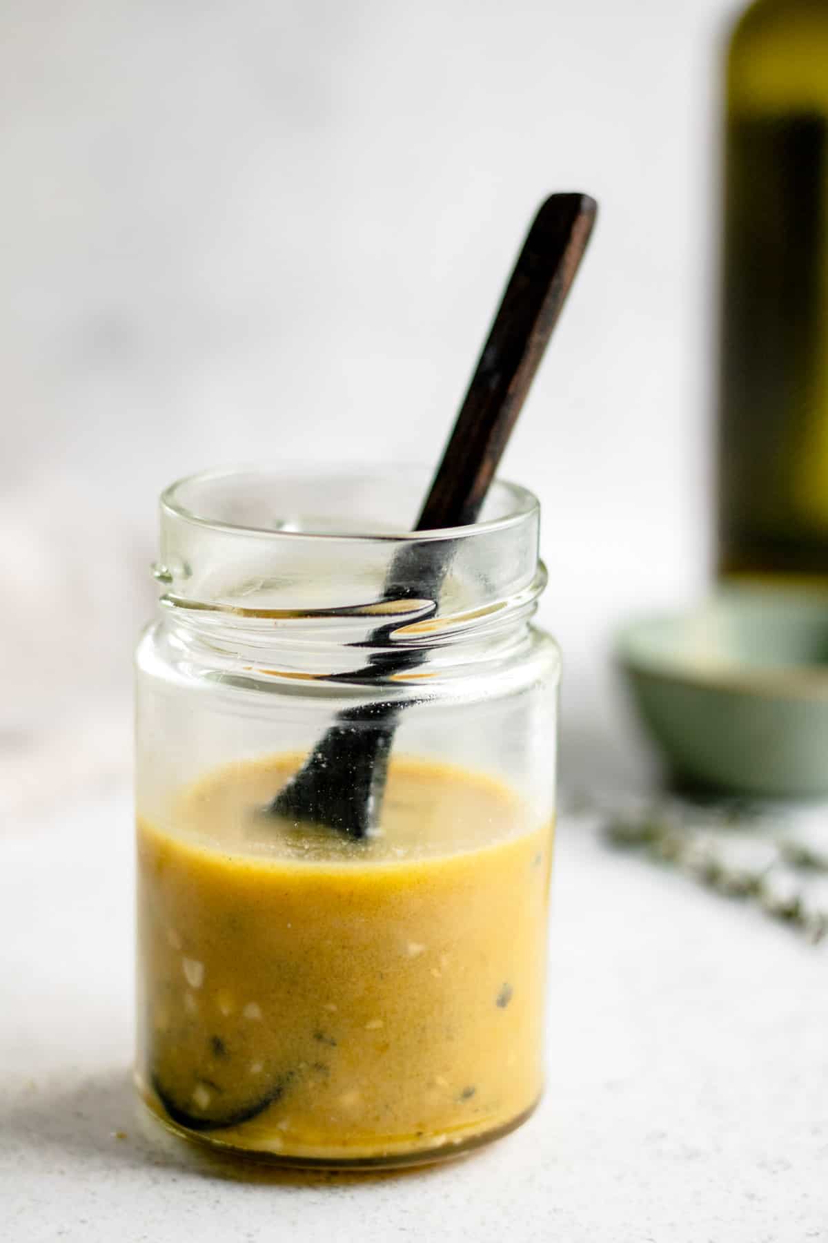 Honey mustard vinaigrette in a glass jar with a wooden spoon and a bottle of extra virgin olive oil in the background.