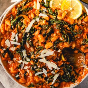 Dish of white beans and kale in tomato sauce garnished with parmesan cheese and lemon.