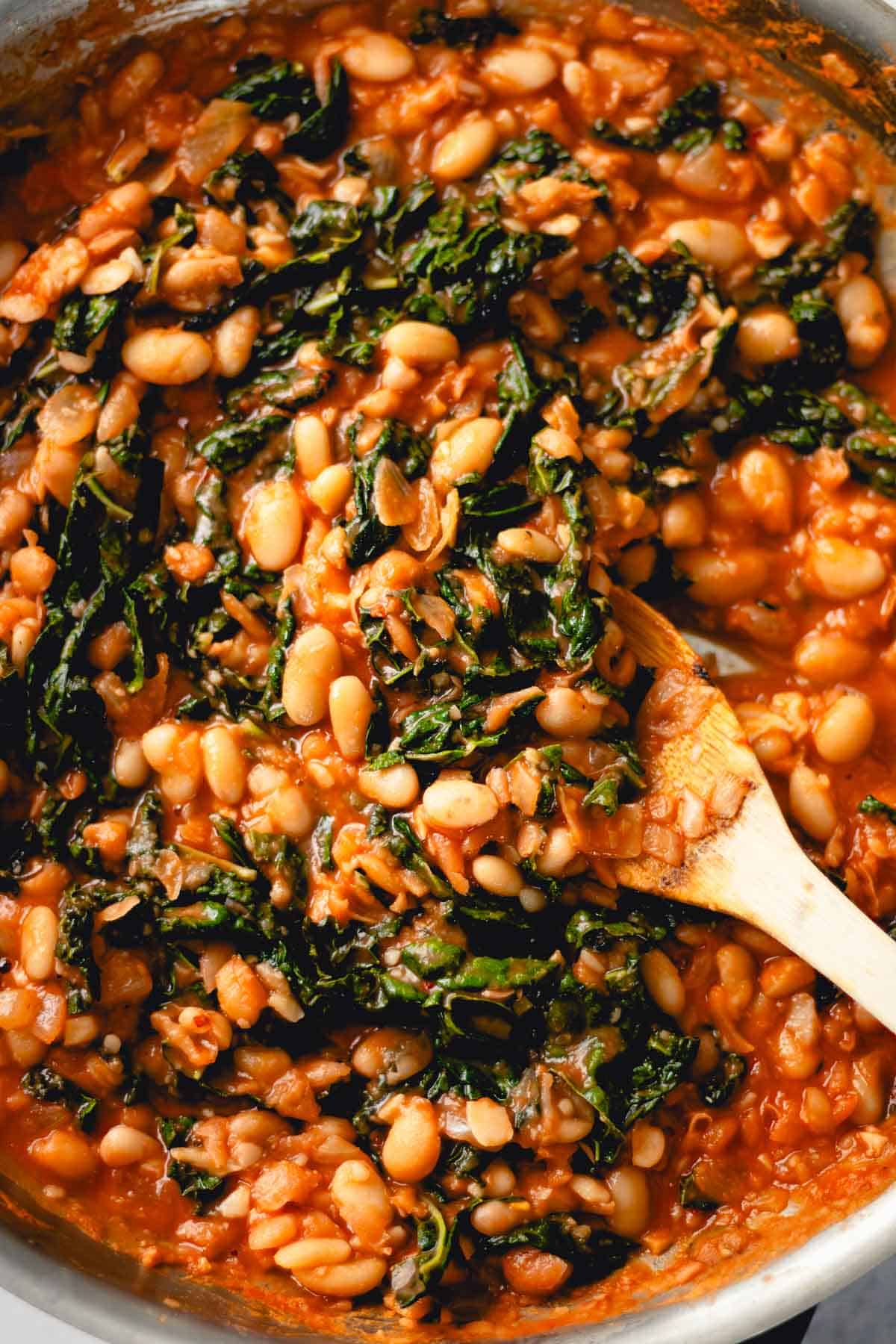 White beans and kale with tomato sauce in a skillet being stirred with a wooden spoon.