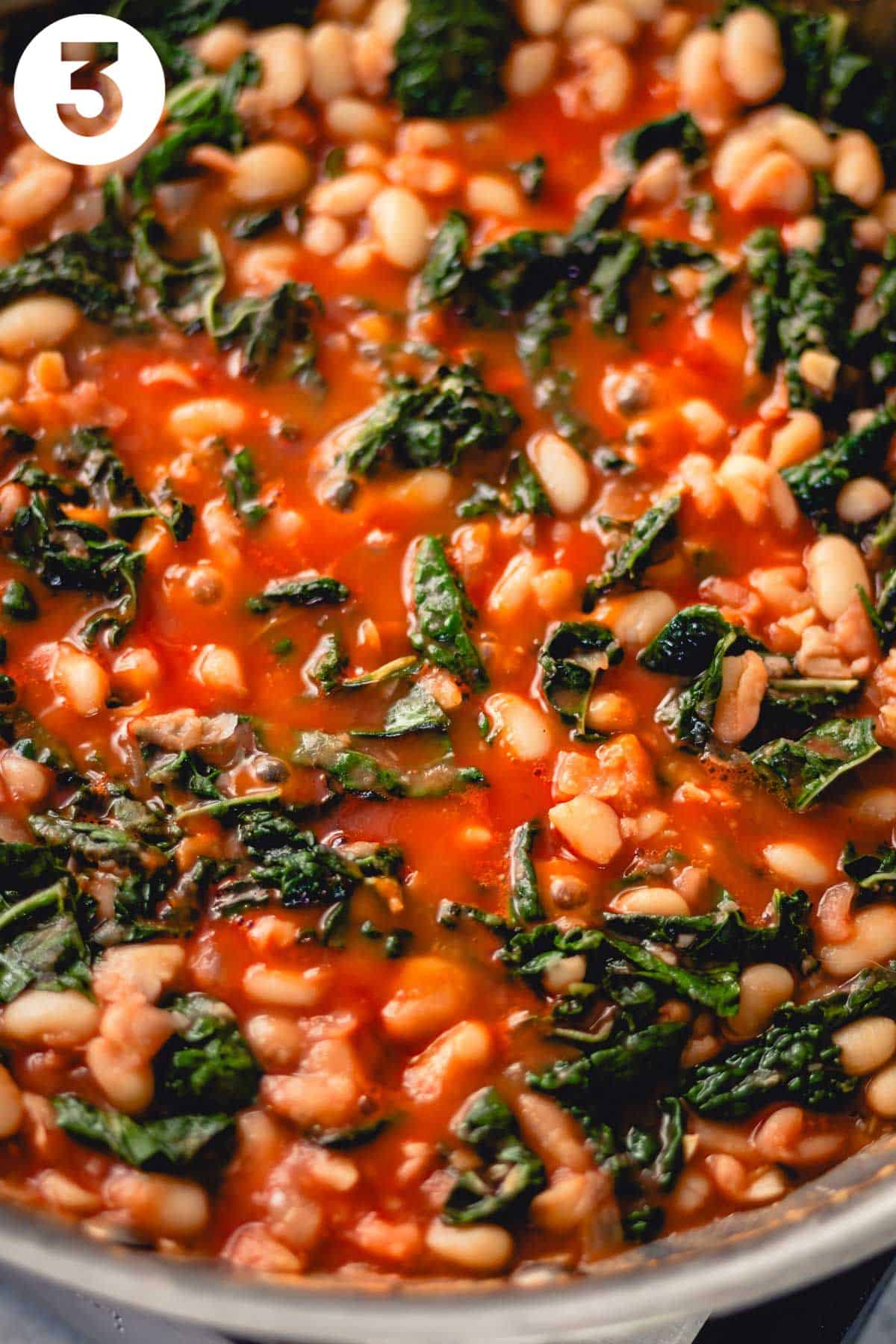 White beans and kale simmering in a tomato-based broth. There is a number "3" in the upper left corner.