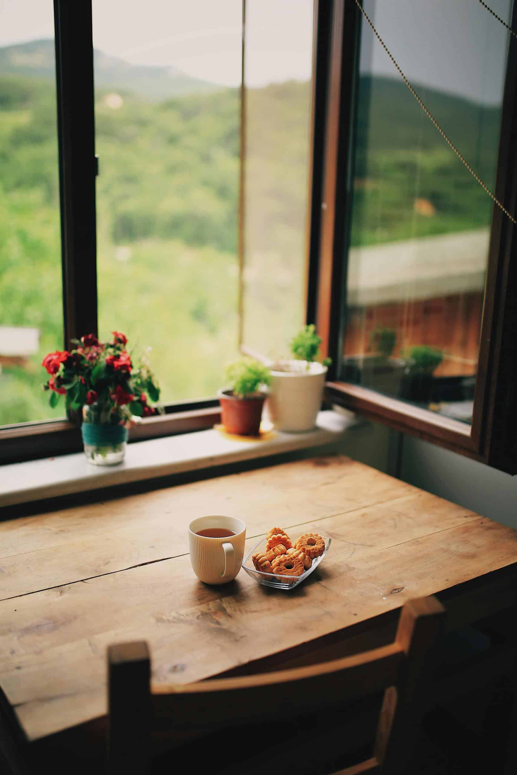 A peaceful morning setup on a wooden table with coffee, pastries, and potted plants by a window.