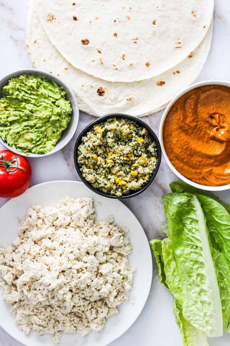 Flatlay photo of burrito ingredients: crumbled tofu, lettuce, quinoa, tomato, avocado, Sofrito sauce, and tortillas.