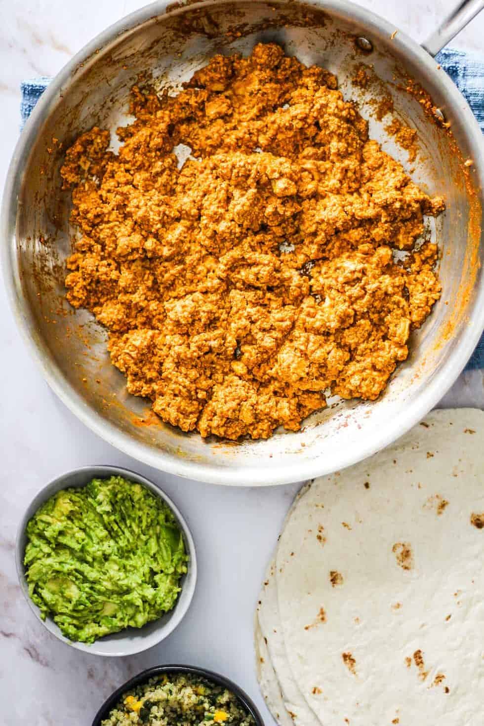 Horizontal image with Sofritas Tofu in a skillet, tortillas, mashed avocado, and quinoa.