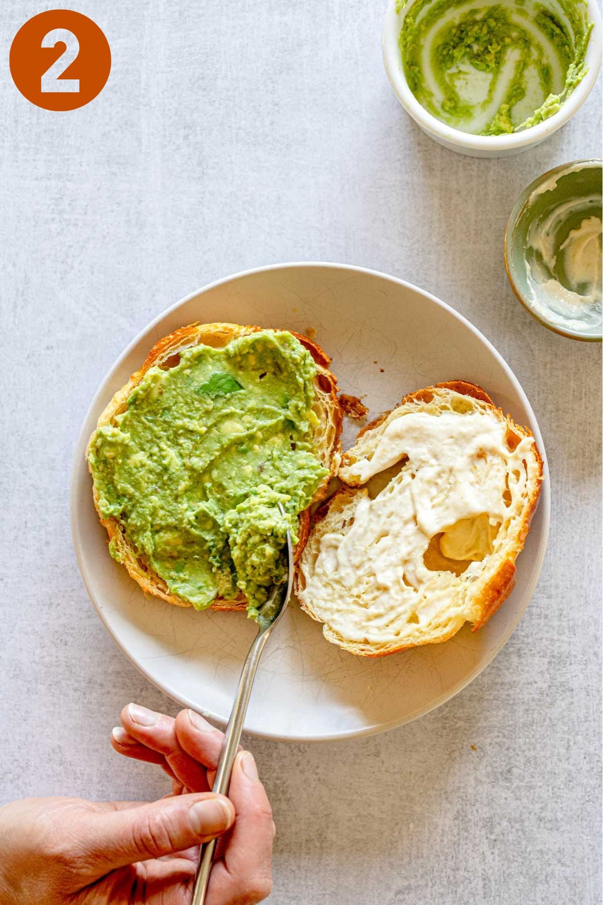 Spreading avocado and aioli on an open croissant.